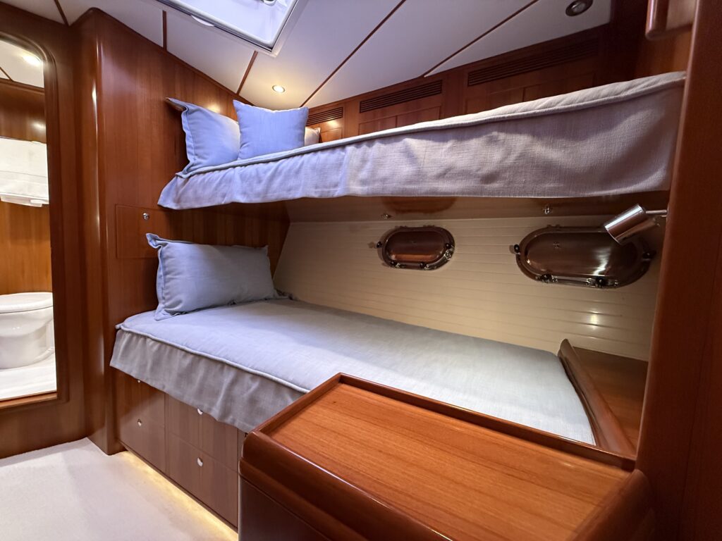 N6416 - AEGEAN MIST - guest stateroom with two bunk berths dressed in light blue linen, brass oval portholes, teak woodwork, built-in drawers, and reading light