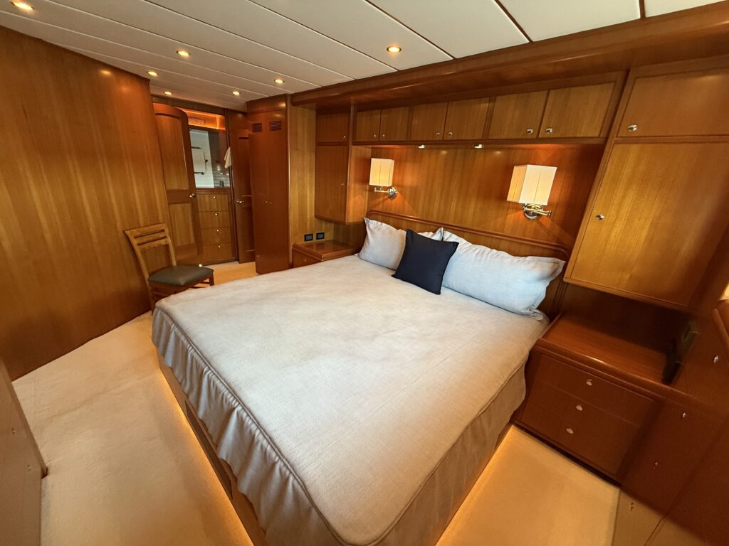 N6416 - AEGEAN MIST - master stateroom with large queen bed, white linens, navy accent pillow, overhead teak cabinetry, flanking reading lamps, and en suite doorway