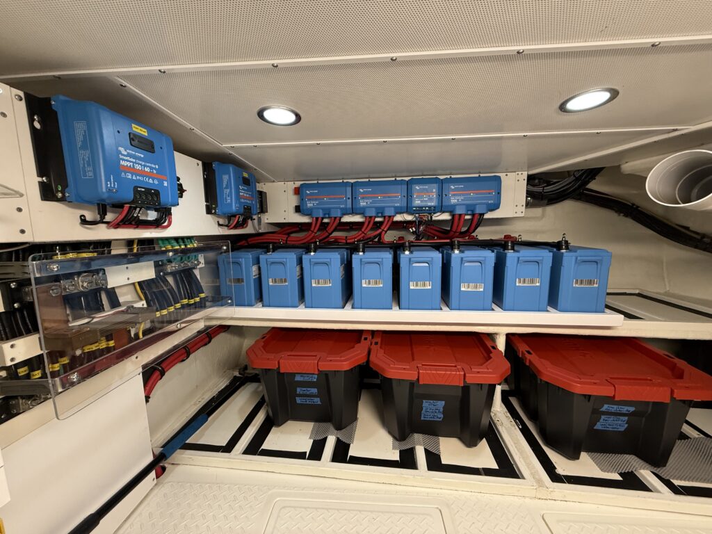 N6416 - AEGEAN MIST - battery bank room showing rows of blue lithium battery cases on shelving, Victron solar charge controllers on the wall, and storage tubs below