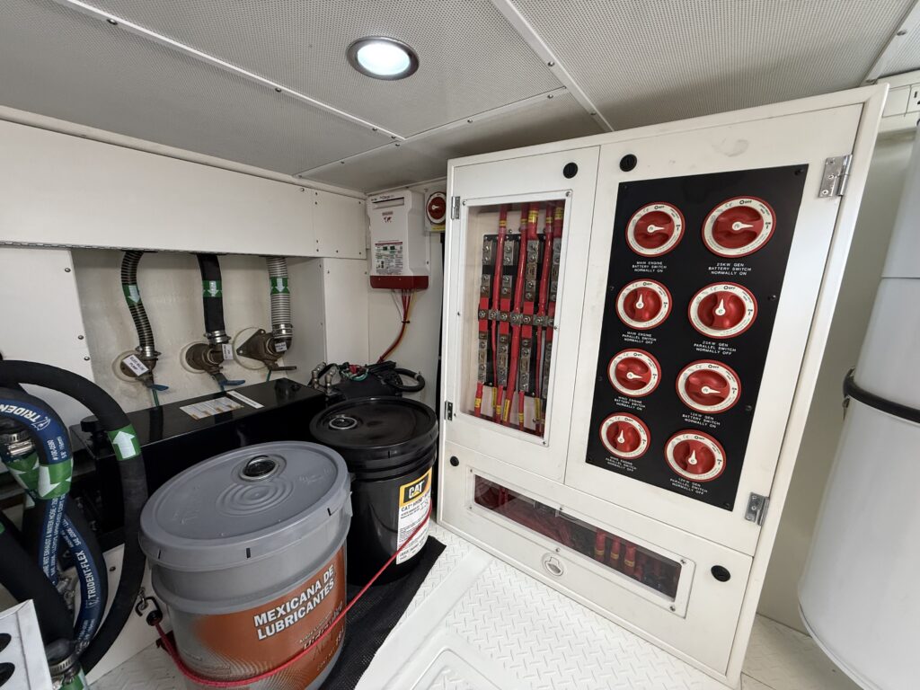 N6416 - AEGEAN MIST - engine room area with hinged cabinet panel showing multiple round pressure gauges, fuel and oil supplies, and bilge hose on deck